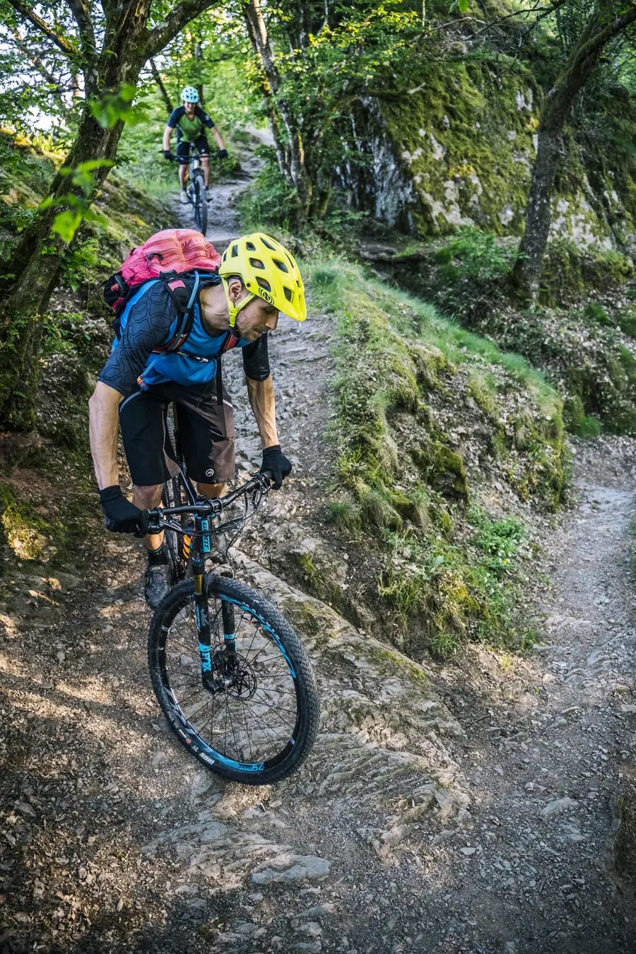 Mountainbiker fährt einen felsigen Waldtrail hinunter, ein zweiter folgt im Hintergrund